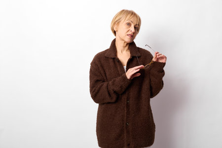 Mature woman in brown coat holding glasses looks thoughtfully to the side on white background....