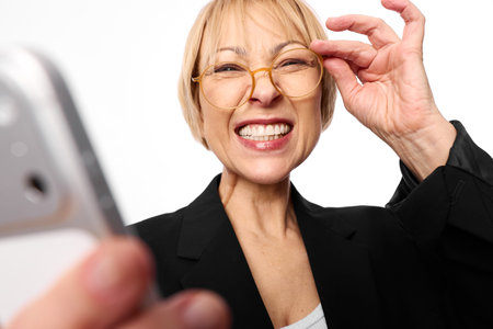 Smiling mature woman with short blonde hair and glasses takes selfie with smartphone on white background.の写真素材