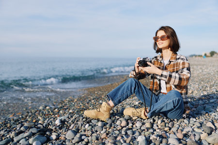 Woman photographer sits on a rocky beach with a handheld camera, enjoying a sunny day by the sea,...