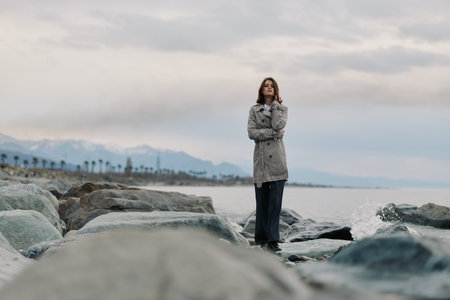A confident woman stands on rocky shore wearing a grey coat and dark pants. The cloudy sky and...