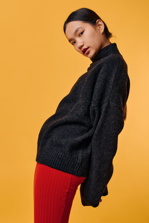 Portrait of young woman wearing black knitted sweater and red pants standing against yellow...