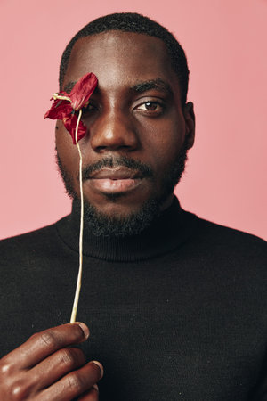 African man with dark skin holding red flower near face in creative conceptual visual style. Portrait of black male model with beard wearing black turtleneck on pink background for artisticの写真素材