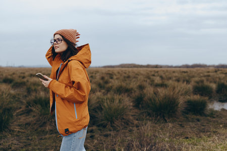 Woman lifestyle in autumn outdoor wearing orange jacket and beanie holding smartphone. She stands in...