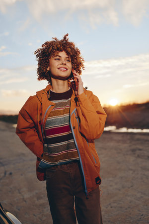 Young woman with curly hair smiles while talking on smartphone outdoors wearing a rainbow sweater...