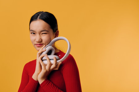 Young woman in red sweater holding white headphones close to her face, smiling with one eye closed...