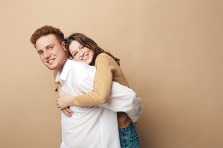 Smiling young couple in casual clothes embraces against beige background. Happy man and woman enjoy...