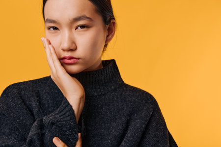 Young woman with hand on face looking serious and thoughtful wearing dark sweater on orange...