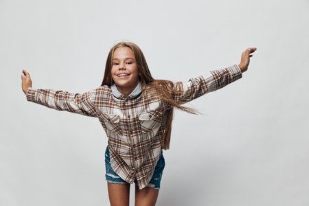 Girl smiling with arms outstretched wearing plaid shirt and denim shorts against white background....