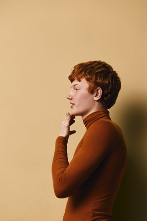 Profile portrait of redhead young man wearing brown turtleneck sweater standing against beige...