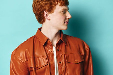 Young man with red hair wears brown leather jacket looking sideways on blue background. Casual...