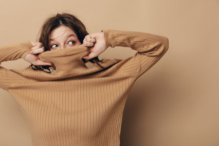 Young woman in brown knitted sweater pulls collar over face, looking sideways with curious expression. Neutral beige background creates warm and cozy atmosphere. Concept of shy or playful mood.の写真素材