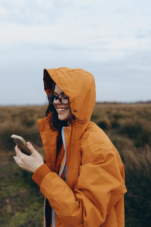 Smiling woman in orange hooded jacket uses smartphone outdoors in natural landscape. Lifestyle concept with casual clothing, technology, and positive mood in overcast weather conditions.の写真素材