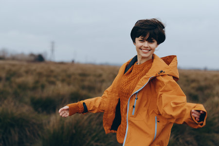 Smiling woman in orange jacket enjoys outdoor lifestyle in autumn field. Happy young female model expresses freedom and joy in natural environment with casual clothing.の写真素材