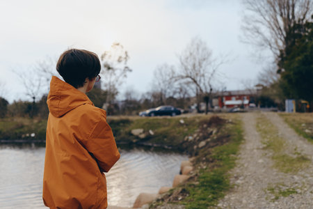Woman lifestyle concept with a person wearing an orange jacket standing near a riverbank on a cloudy day. The scene shows nature, autumn, and thoughtful mood outdoors.の写真素材