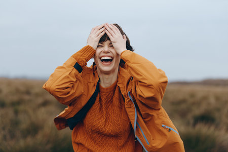 Happy woman lifestyle outdoors wearing orange jacket and sweater laughs joyfully with hands on head....