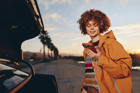 Young woman smiling outdoors near car trunk at sunset, wearing sweater and orange jacket, holding...