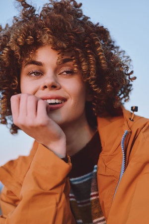 A smiling woman with curly hair wears a rainbow striped sweater and an orange jacket while sitting...