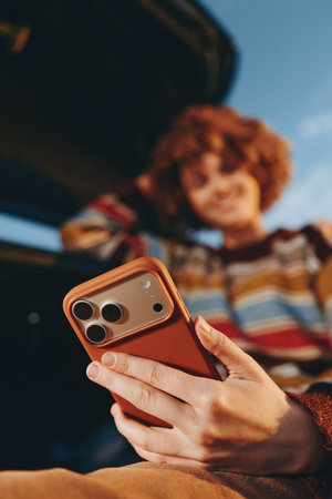 Woman with curly hair wearing rainbow sweater smiling while holding smartphone outdoors near car...