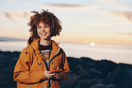 Smiling woman with curly hair wearing a rainbow sweater and orange jacket holding a smartphone near...