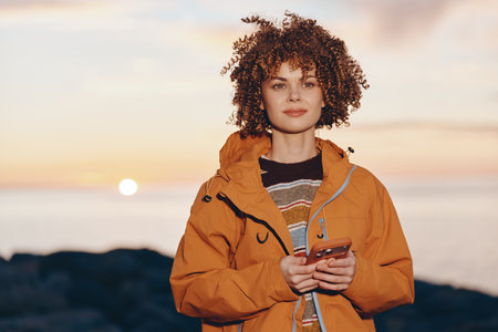 Young woman with curly hair wearing orange jacket and rainbow sweater, holding smartphone, standing...