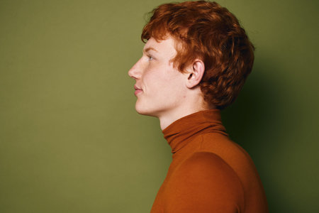 Profile portrait of young man with red hair wearing orange turtleneck standing against green...