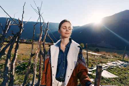 Woman portrait in mountains with sunlight and nature, peaceful expression with eyes closed, wearing warm jacket in rural outdoor scene, relaxation, fresh air and mindful moment.の写真素材