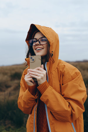 Smiling woman in orange jacket and glasses holds smartphone outdoors in natural setting. Lifestyle...