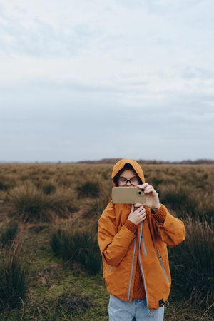 Young woman lifestyle outdoors takes photo with smartphone in autumn jacket on moor landscape under...
