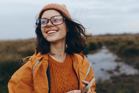 Smiling woman in orange jacket and sweater enjoys outdoor lifestyle with glasses and hat. Natural...
