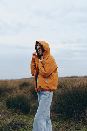 Young woman lifestyle outdoors wearing orange hooded jacket and sunglasses stands in grassy field...