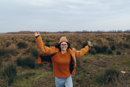 Happy woman in orange sweater and jacket enjoys outdoor lifestyle in open field, smiling with raised...