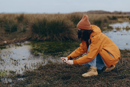 Young woman in orange jacket and beanie crouches near wetland, taking photo with smartphone. Outdoor...