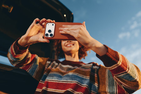 Woman smiling outdoors wearing a rainbow sweater taking a selfie with smartphone near car under...