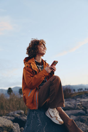 Young woman with curly hair wearing an orange jacket and brown pants sits on a rock outdoors using a...