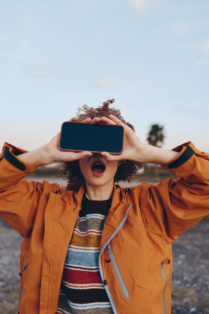 Young woman outdoors holding smartphone horizontally in front of her face, wearing colorful striped sweater and orange jacket, playful expression, natural light, casual lifestyle momentの写真素材