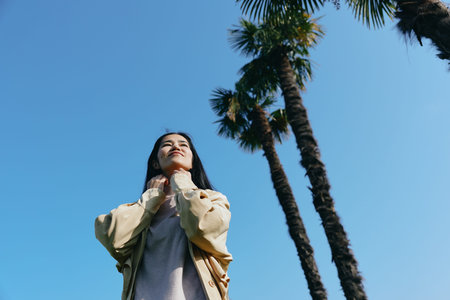 Skyward view of a woman outdoors beneath a bright blue sky, tall palms beside her, calm gaze upward,...
