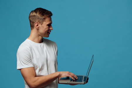 young caucasian man working on laptop wearing white t-shirt standing side view on blue background digital nomad remote work conceptの写真素材