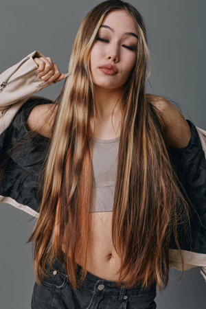 Young woman with long straight hair wearing a crop top and jacket posing against gray background...