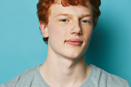 Young man with red hair and fair skin wearing a gray shirt poses against blue background looking...