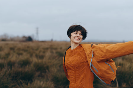 Happy woman lifestyle outdoors wearing orange sweater and jacket spreads arms in open field under...