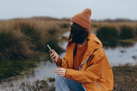 Woman in orange jacket and knit hat uses smartphone outdoors near pond. Autumn nature background...