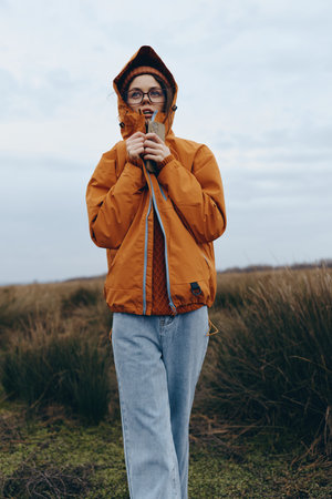 Young woman lifestyle in orange jacket with hood stands outdoors in nature field under cloudy sky....