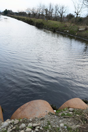 Calm river flows through a natural landscape with leafless trees and grassy banks under an overcast...