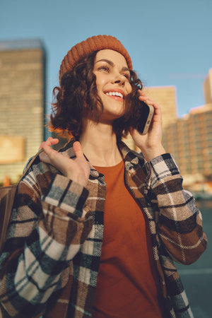 Woman on phone smile in city street wearing beanie and plaid jacket, sunlight and golden hour glow;...