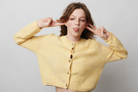 Young woman in yellow knitted sweater makes playful face with tongue out and fingers near ears....
