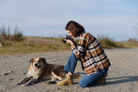 Photographer kneeling in a sandy outdoor setting, capturing a playful dog with a compact camera,...