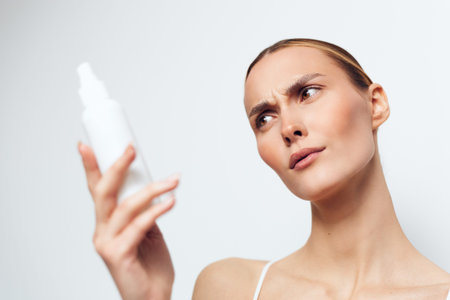 Young woman with clear skin holding white cosmetic bottle close to face, examining product...