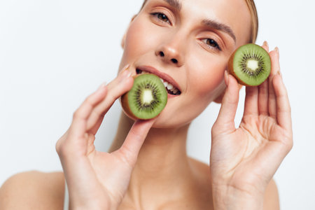 A smiling woman holds two halves of a fresh kiwi fruit near her face against a white background. The...