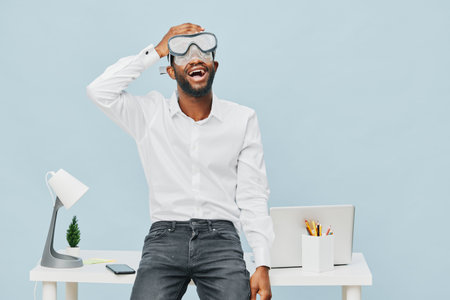 Smiling man with beard wearing protective goggles and white shirt standing in modern office with laptop and desk, enjoying work or fun moment, full body, casual style, bright background, office lifestyle concept.の写真素材
