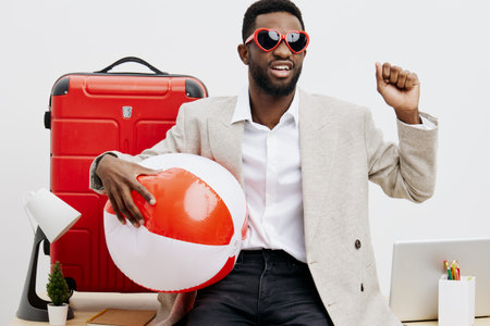 Young smiling man with dark skin, wearing beige blazer, holding an orange and white beach ball, sitting at desk with travel suitcase, computer and stationery on white background. People lifestyle conceptの写真素材